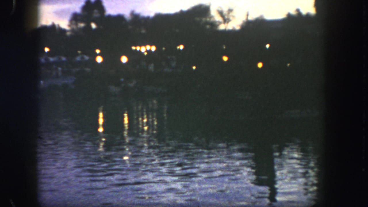 1962 Disneyland 8mm frame showing dim shoreline lights reflected on a lake at twilight, with trees in silhouette.
