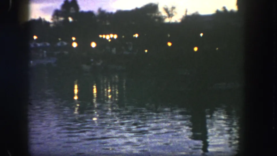 1962 Disneyland 8mm frame showing dim shoreline lights reflected on a lake at twilight, with trees in silhouette.