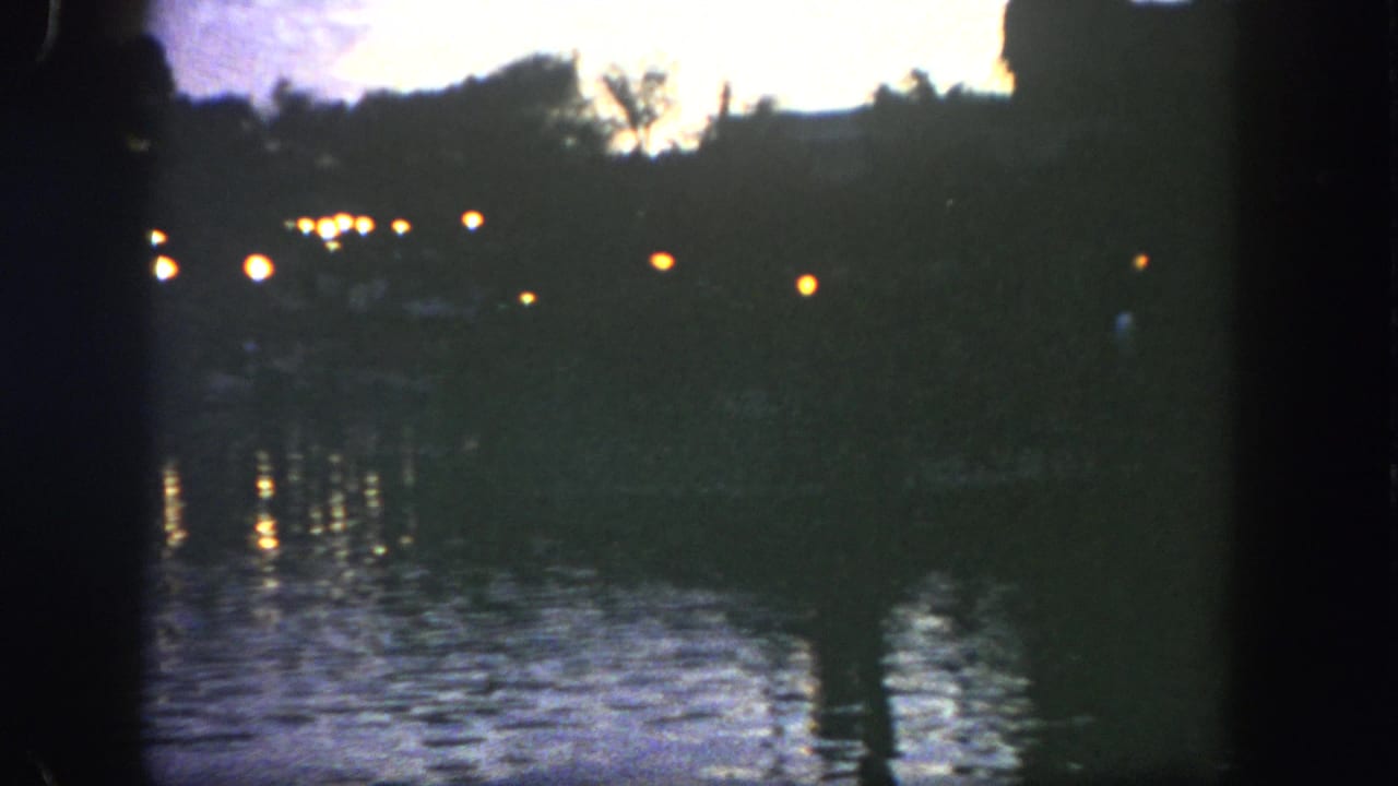 1962 Disneyland 8mm view of a dark lakeside shoreline with warm lamps and wavering reflections on the water.