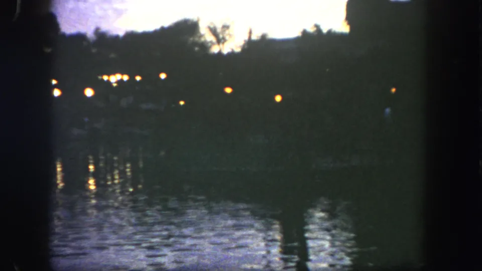 1962 Disneyland 8mm view of a dark lakeside shoreline with warm lamps and wavering reflections on the water.
