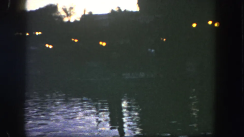 1962 Disneyland 8mm frame with scattered lights beyond a dark lake, softened by low-light exposure and grain.