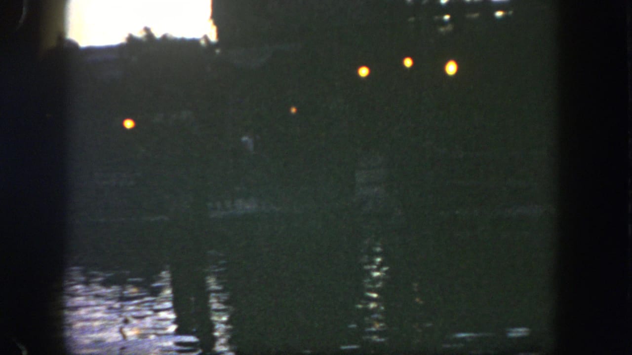 Dim river water at dusk with small warm lights reflected near dark silhouettes.