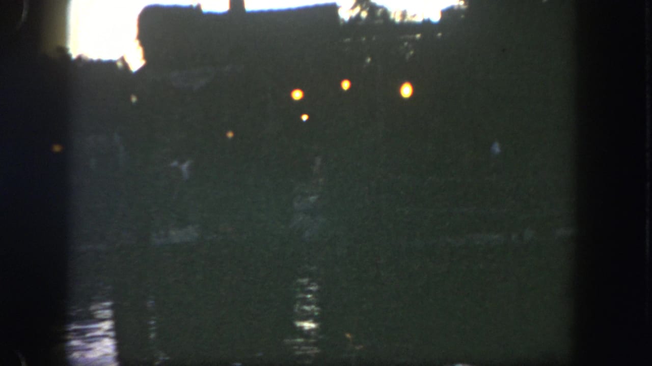 1962 Disneyland riverfront frame with silhouetted structures or trees, warm lamps, and low-light water reflections.