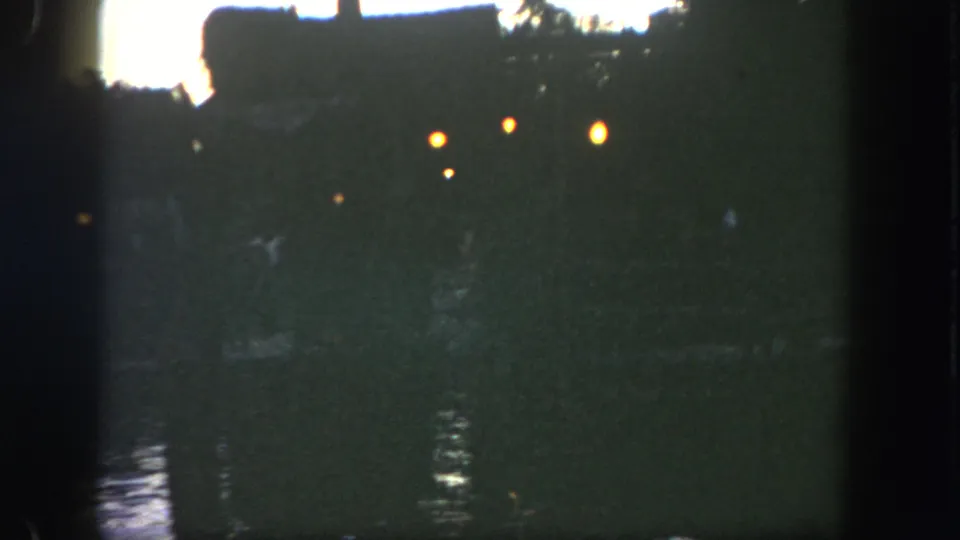 1962 Disneyland riverfront frame with silhouetted structures or trees, warm lamps, and low-light water reflections.