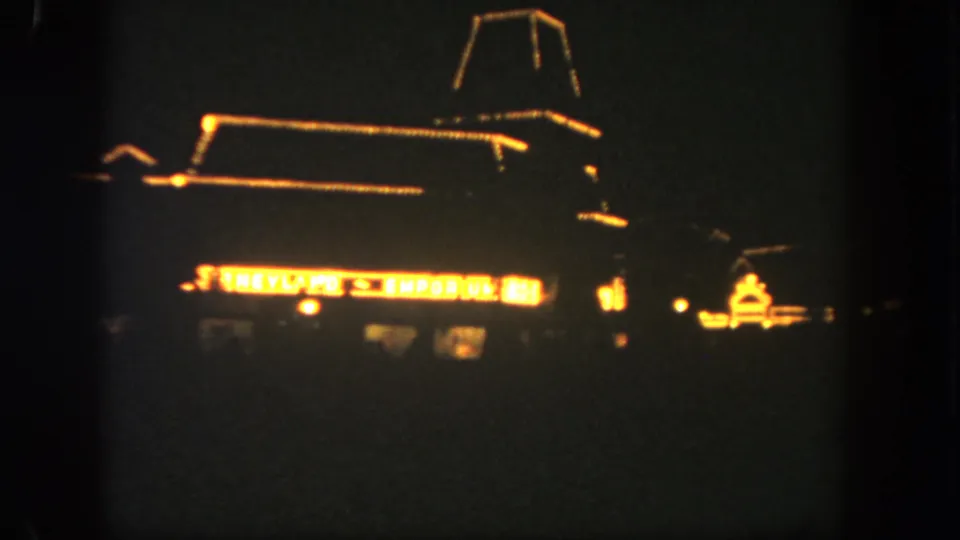 1962 Disneyland nighttime home-movie frame showing the Emporium facade outlined in warm lights with a bright sign below.