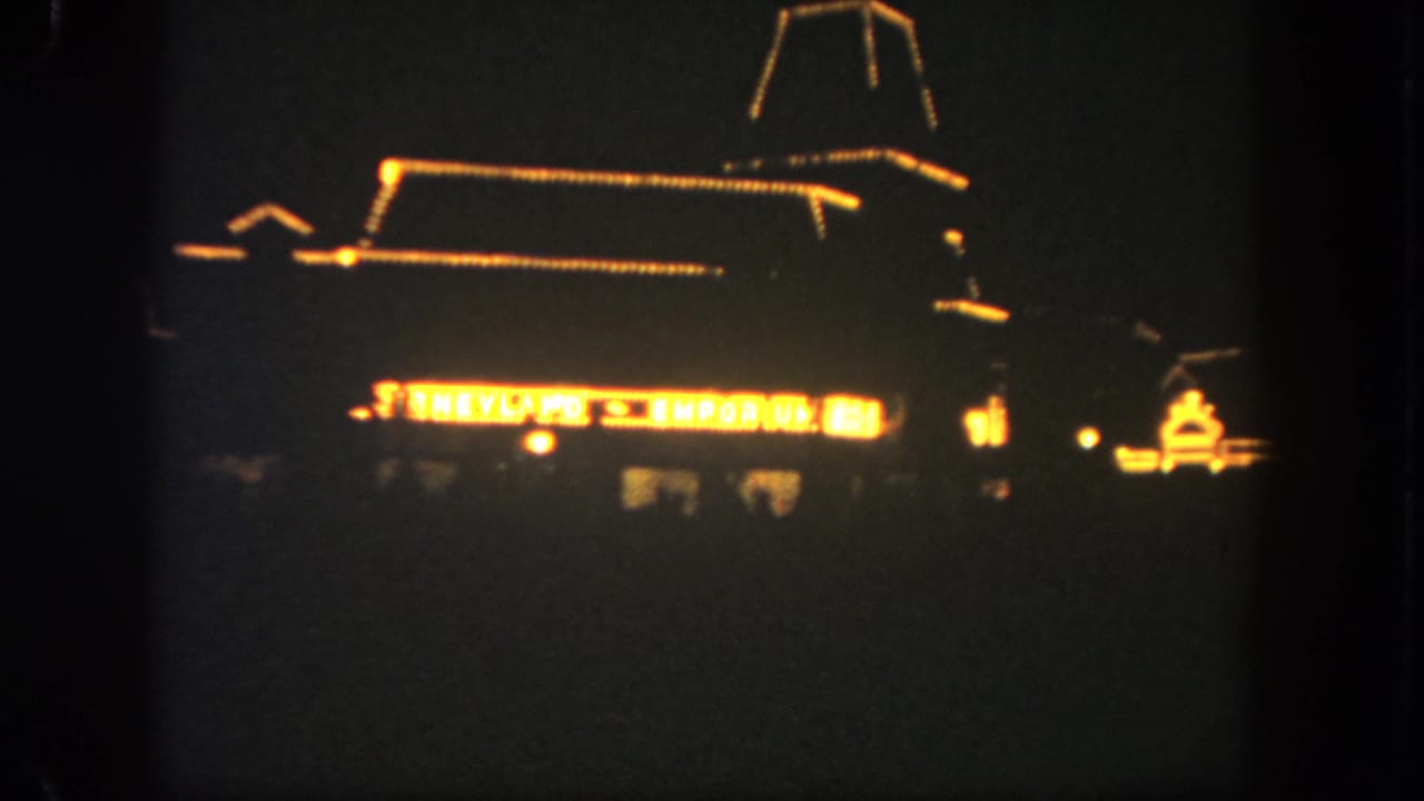 Visitor-shot 8mm view of the Disneyland Emporium area at night, with warm outline lights and a bright sign visible.