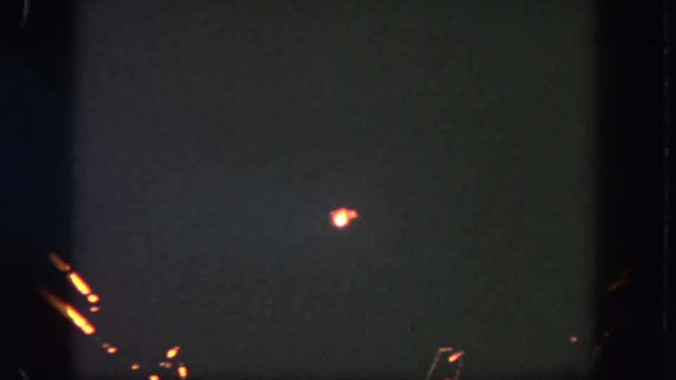 The 1962 8mm frame shows a small orange burst near the lower center, with dark exposure, grain, and edge shadows.