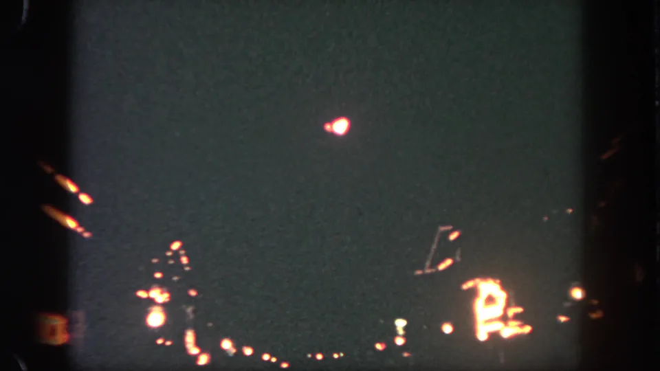 Nighttime Disneyland frame with a red-white pyrotechnic burst above soft distant lights and dark 8mm frame edges.