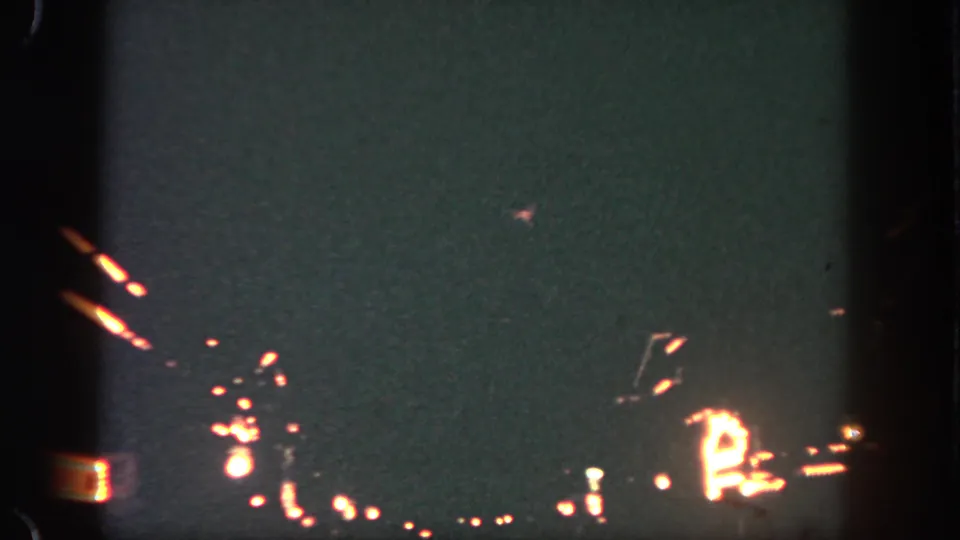 Low-light 8mm frame from Disneyland showing scattered ground lights and faint pyrotechnic traces in the dark sky.