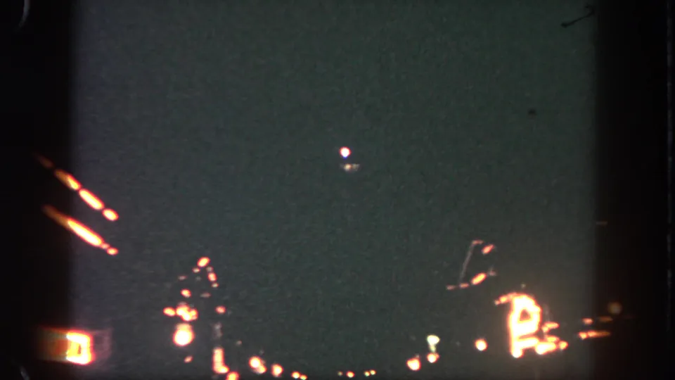 1962 Disneyland fireworks frame with a small white burst above warm orange trails in grainy 8mm footage.