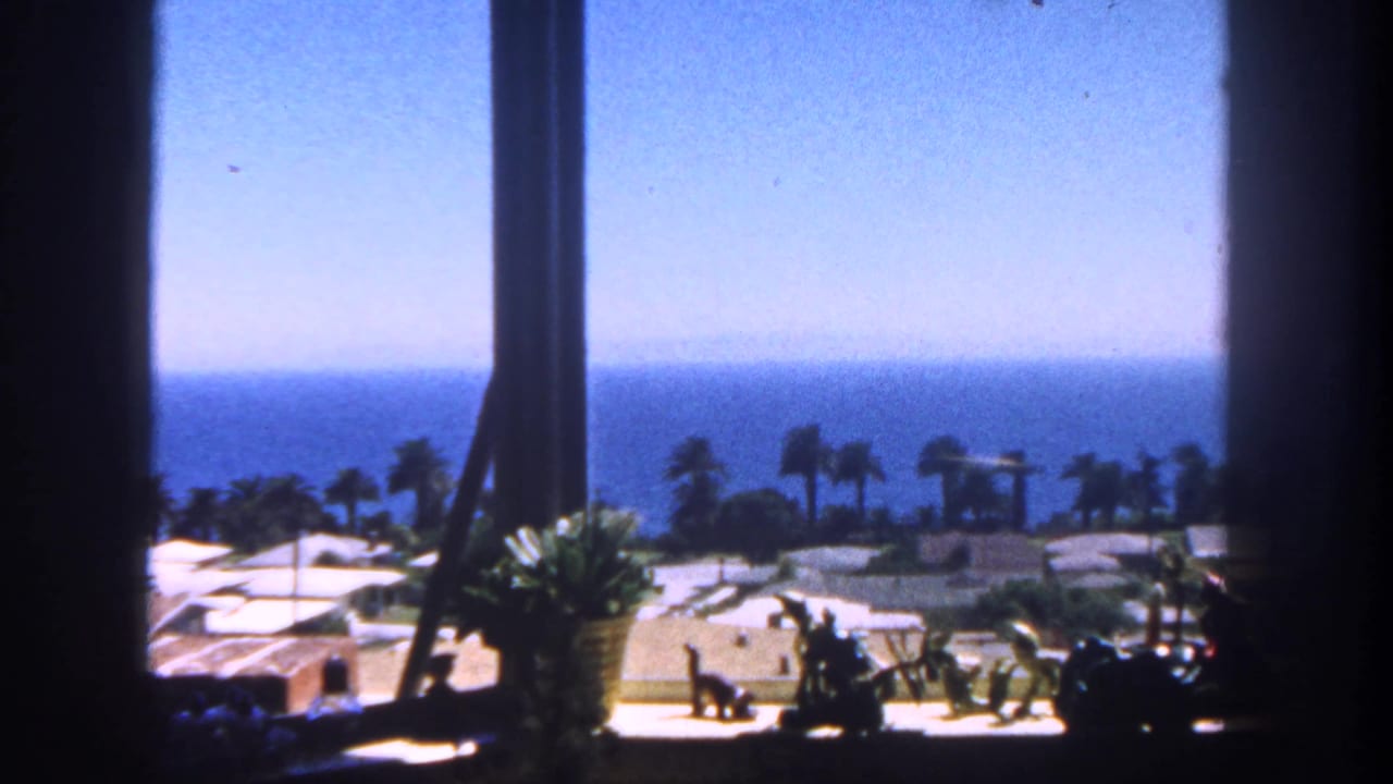 1962 San Diego 8mm frame looking from a shaded window toward palm trees, low coastal buildings, beach ground, and the ocean horizon.