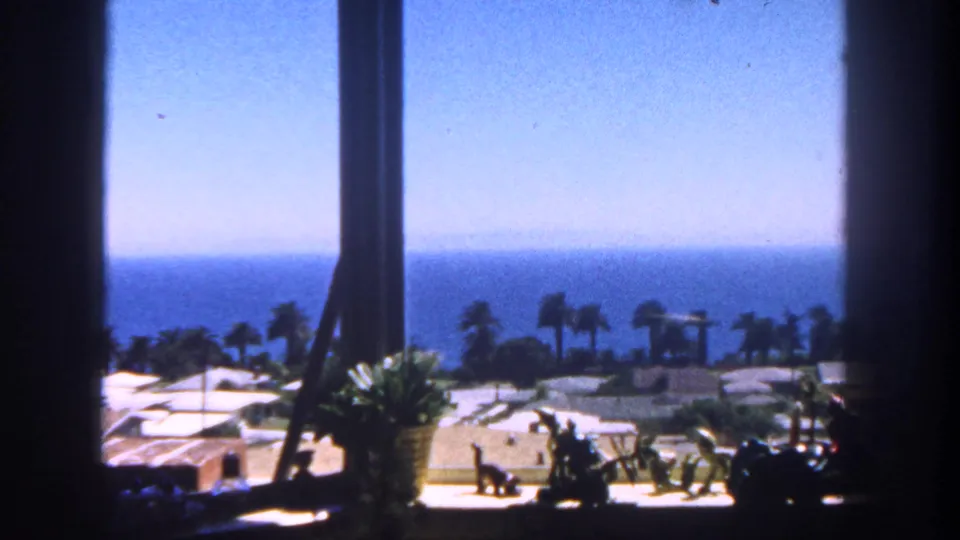 1962 San Diego 8mm frame looking from a shaded window toward palm trees, low coastal buildings, beach ground, and the ocean horizon.