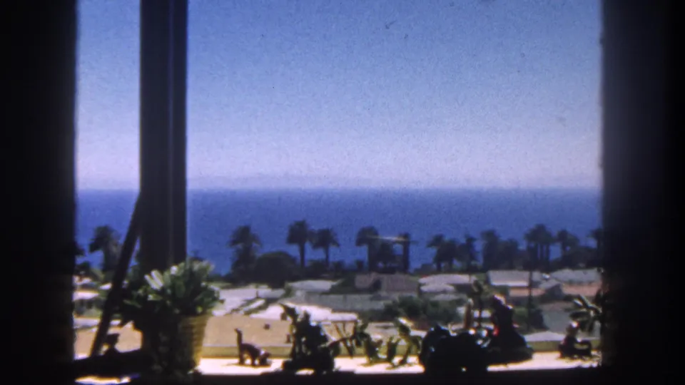 Soft 1962 San Diego home-movie frame from a shaded window, showing palms, coastal buildings, beach area, and ocean under a clear sky.