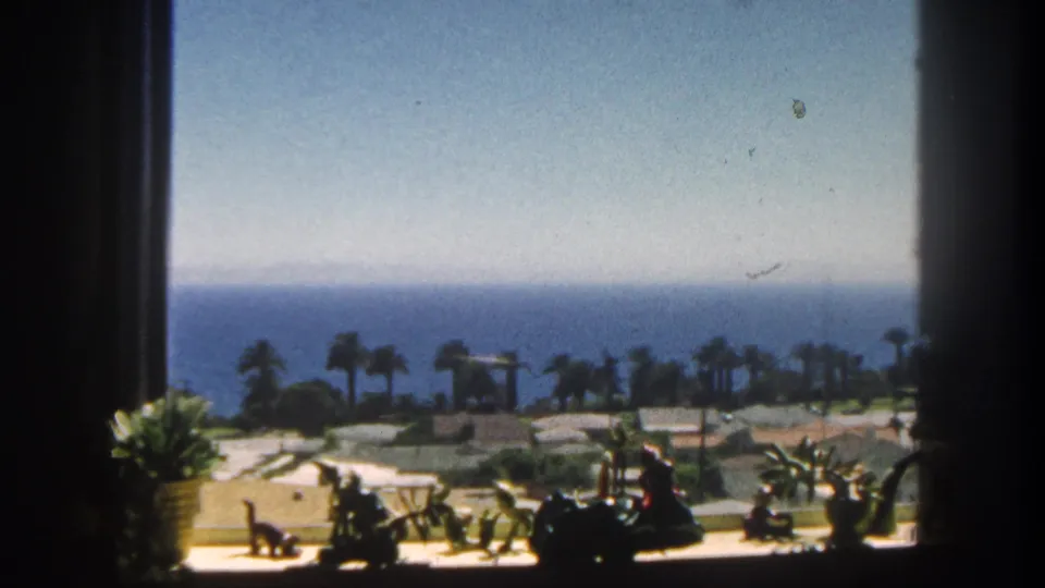1962 San Diego 8mm frame showing a shaded window foreground and a bright palm-lined coastal resort area facing the ocean.