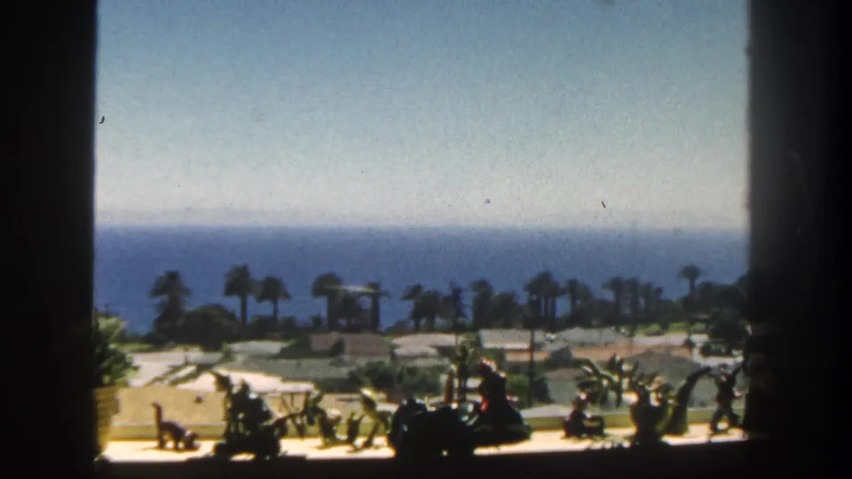 1962 San Diego home-movie frame looking past dark window bars, small sill objects, palms, and low coastal grounds toward the ocean.