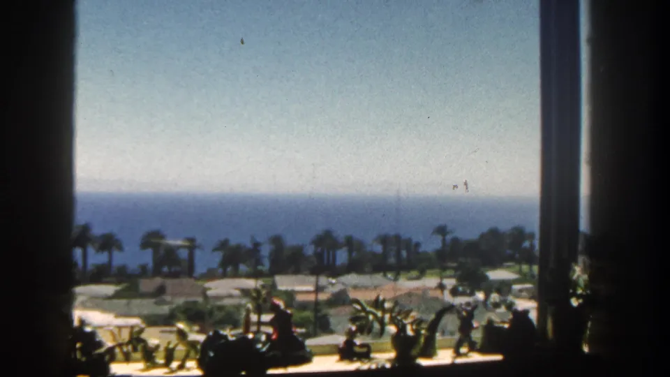 Soft 8mm frame from San Diego in 1962 showing palms, low grounds, and the ocean horizon beyond a dark interior window frame.