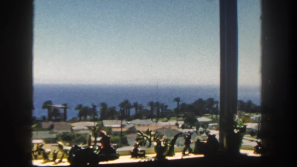 Final 1962 San Diego frame with window bars, small sill decorations, palm trees, low coastal grounds, and the Pacific horizon in bright sun.