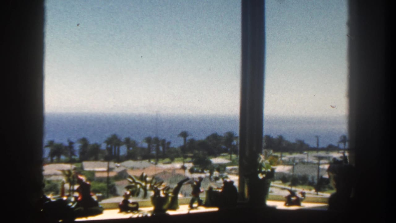 1962 San Diego home-movie frame showing a sunlit coastal view through window mullions, with sill plants, palms, rooftops, and ocean beyond.