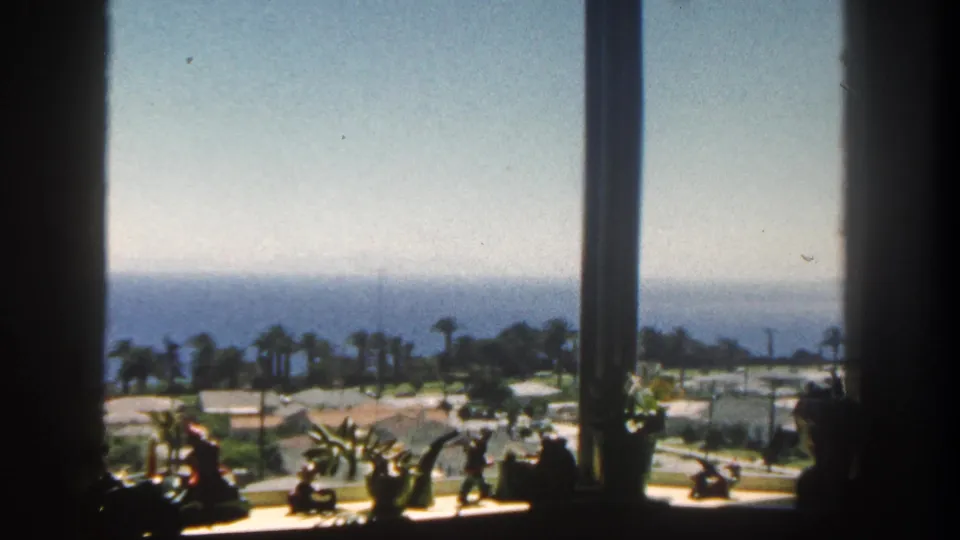 1962 San Diego home-movie frame showing a sunlit coastal view through window mullions, with sill plants, palms, rooftops, and ocean beyond.