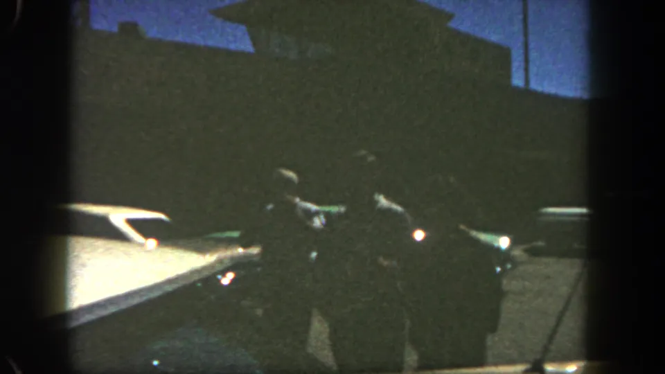 1962 San Diego home-movie frame with three silhouetted figures walking across a parking lot beside cars at dusk.