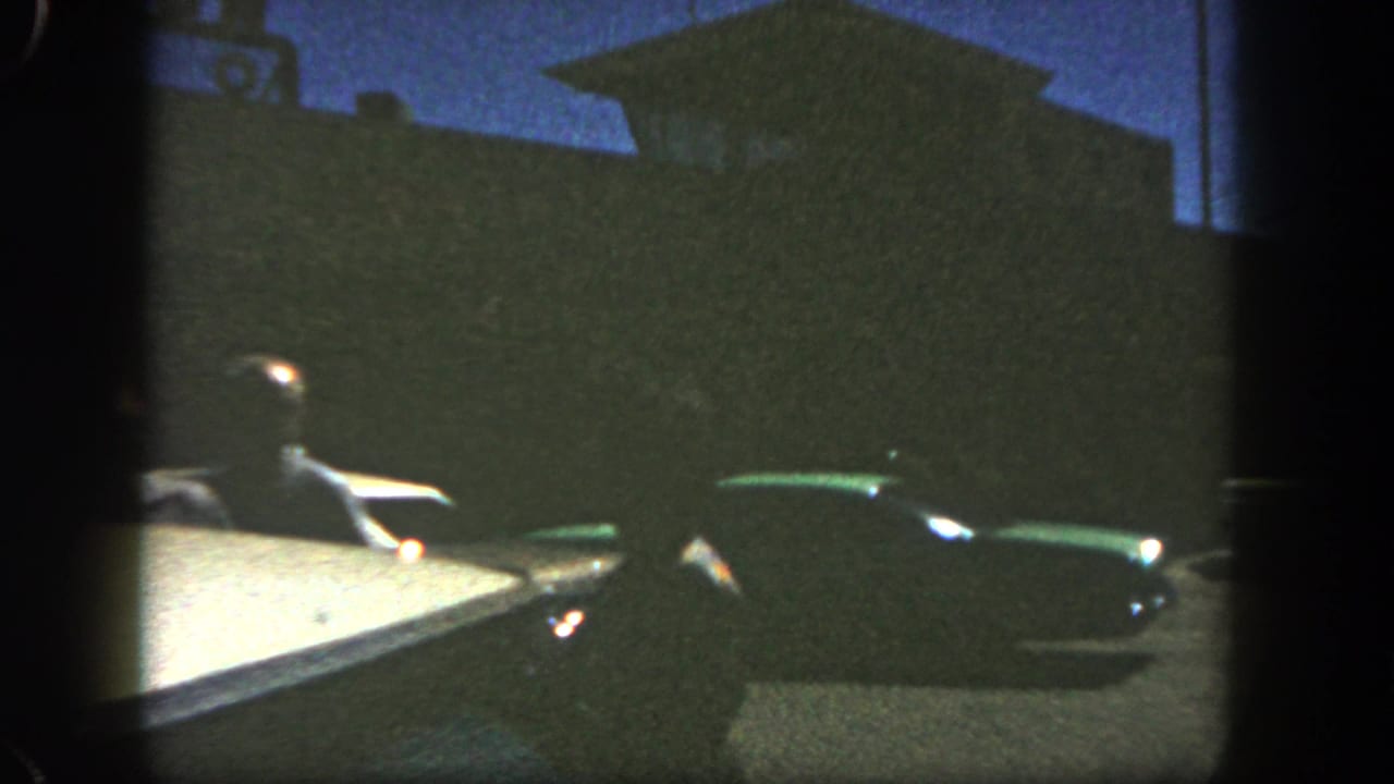 1962 San Diego home-movie frame showing a driver seen from a vehicle, with a dark green classic car and silhouetted roadside building beyond.