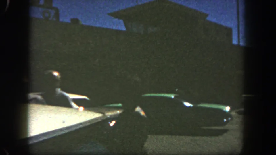 1962 San Diego home-movie frame showing a driver seen from a vehicle, with a dark green classic car and silhouetted roadside building beyond.