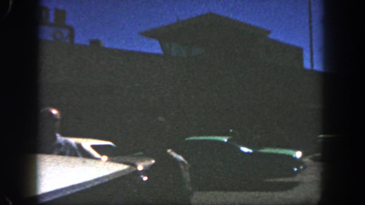 1962 San Diego frame with a driver at left, a dark green mid-century car, and a shadowed roadside structure under blue dusk light.
