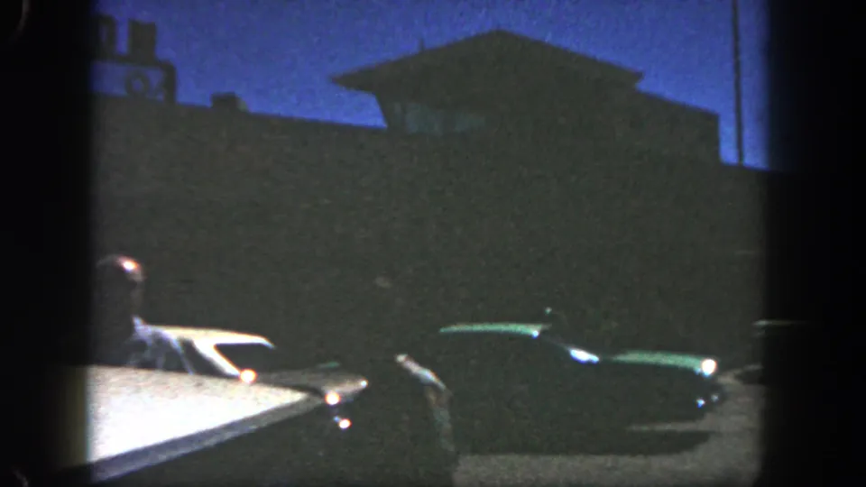 1962 San Diego frame with a driver at left, a dark green mid-century car, and a shadowed roadside structure under blue dusk light.