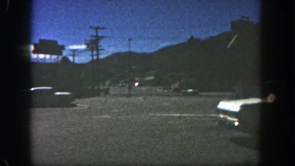 1962 San Diego frame showing a dim roadside view with classic cars, utility poles, distant hills, and a billboard-like structure in the background.