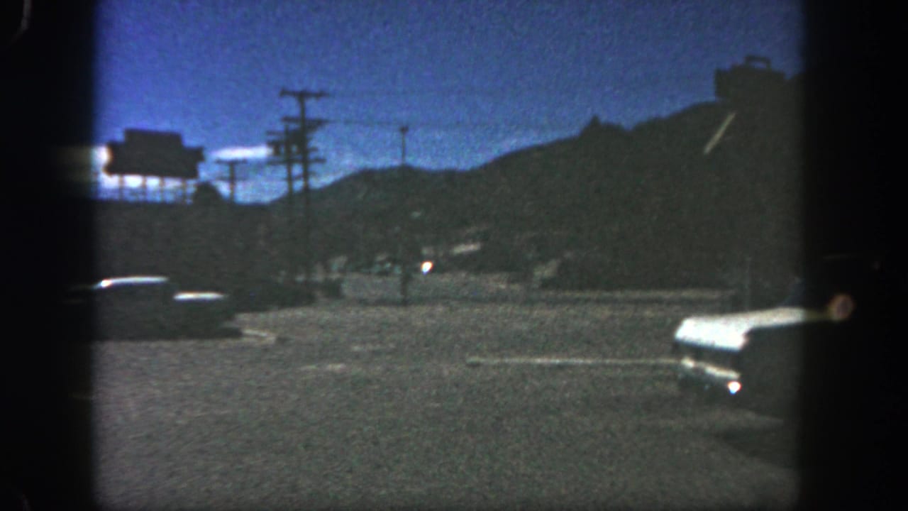 1962 San Diego home-movie frame showing parked cars, utility poles, and dark hills under a blue dusk sky.