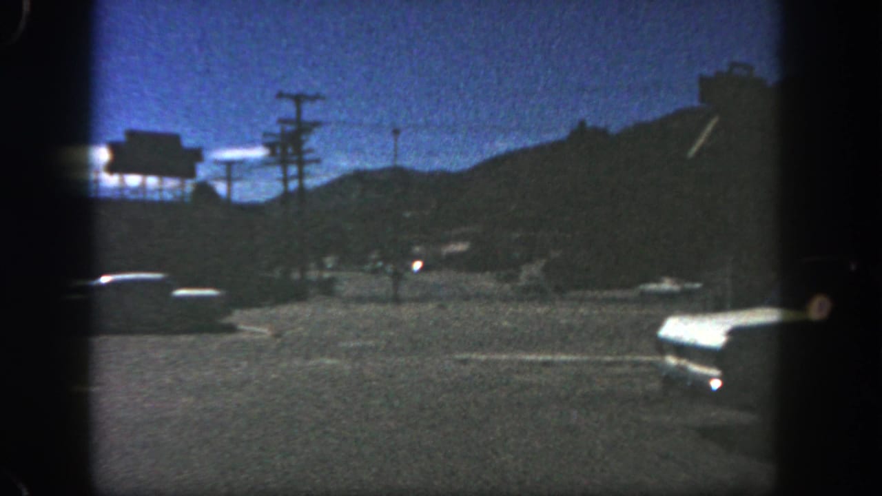 Grainy 8mm view of a San Diego roadside at dusk, with classic cars and utility poles against dark hills.