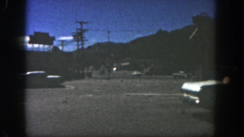 Low-light 8mm frame of a 1962 San Diego roadside, with cars and silhouetted hills visible through heavy grain.