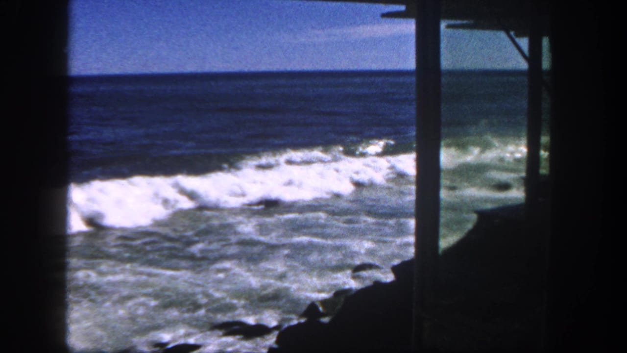 1962 San Diego coastal view with white surf, blue ocean horizon, and dark building posts framing the shoreline.