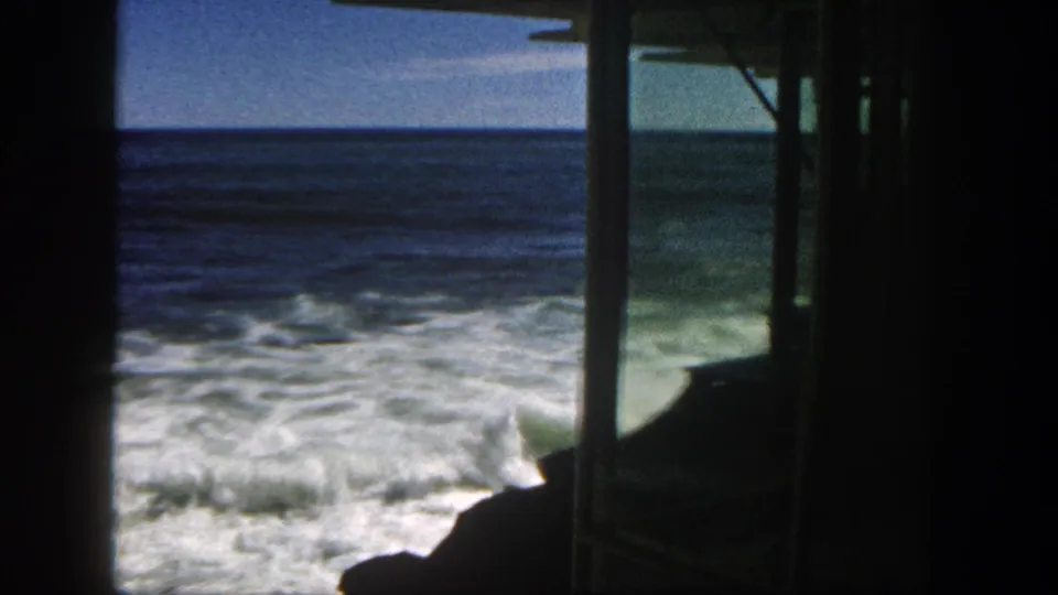 San Diego surf in 1962 rolls past dark rocks, with shaded structural posts enclosing the home-movie view.