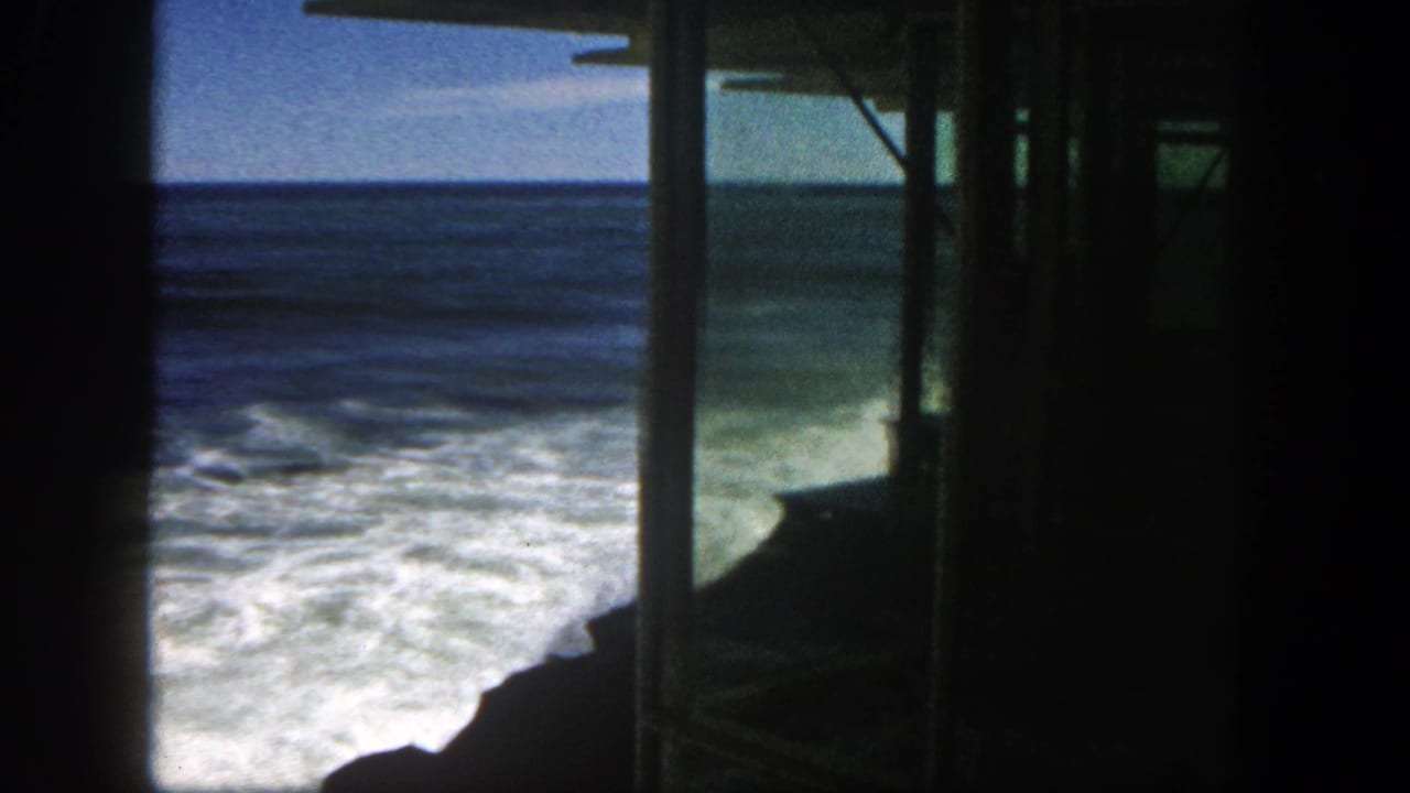 1962 San Diego coastal surf viewed from inside a dark pier-like or beachside structure, with waves breaking near rocks.