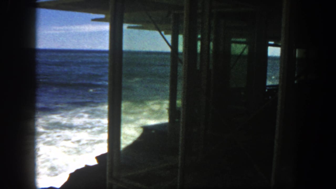 Sunlit San Diego ocean waves and rocky shoreline framed by dark structural posts in 1962 8mm home-movie footage.