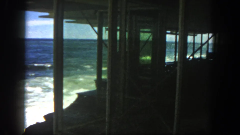 A 1962 home-movie view pans across San Diego surf, with posts and cross-bracing silhouetted against the ocean.