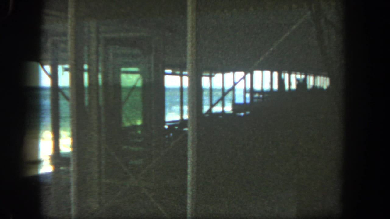Shadowed 8mm view beneath a San Diego pier, with vertical supports silhouetted against surf and blue water.