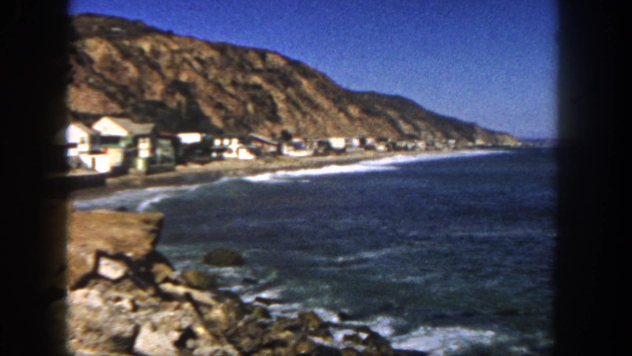 Coastal view with beach houses below a rocky hillside and waves along the shore.