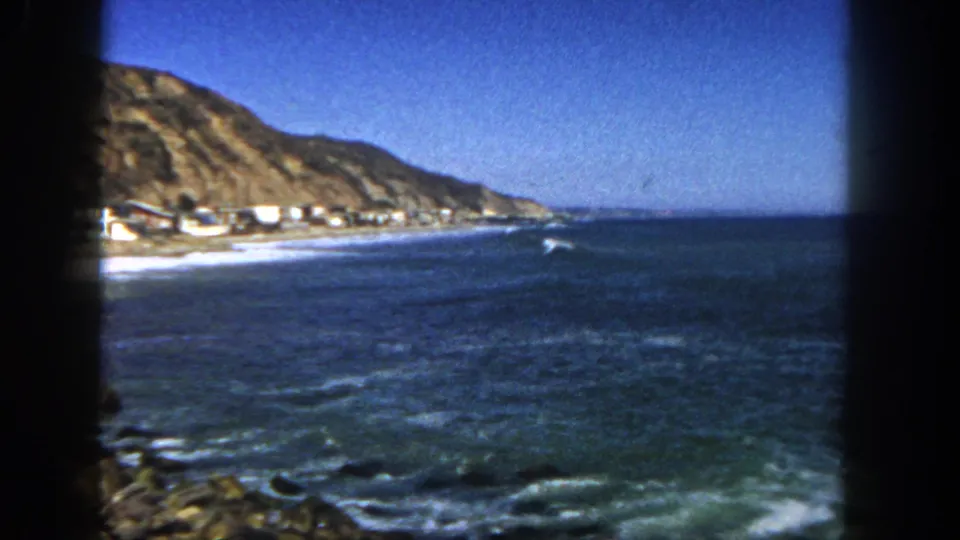 1962 San Diego coastal frame with blue ocean, distant beach houses, rocky shore, hillside, and a bird crossing above the water.
