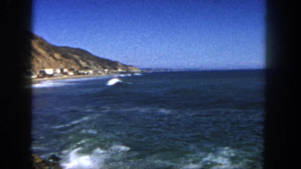 San Diego coast in 1962 with hillside, shoreline homes, deep blue ocean, white surf, and visible 8mm frame edges.