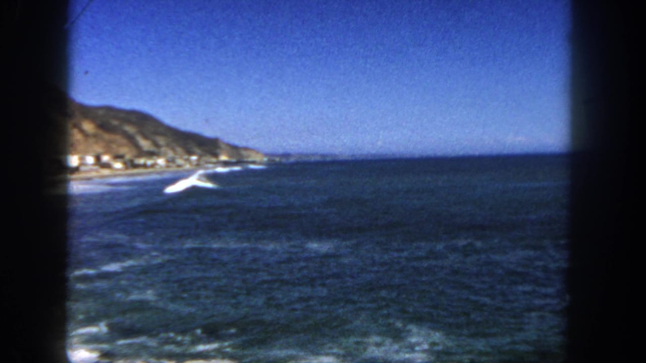Ocean waves below a distant cliff with blue sky and dark film borders.