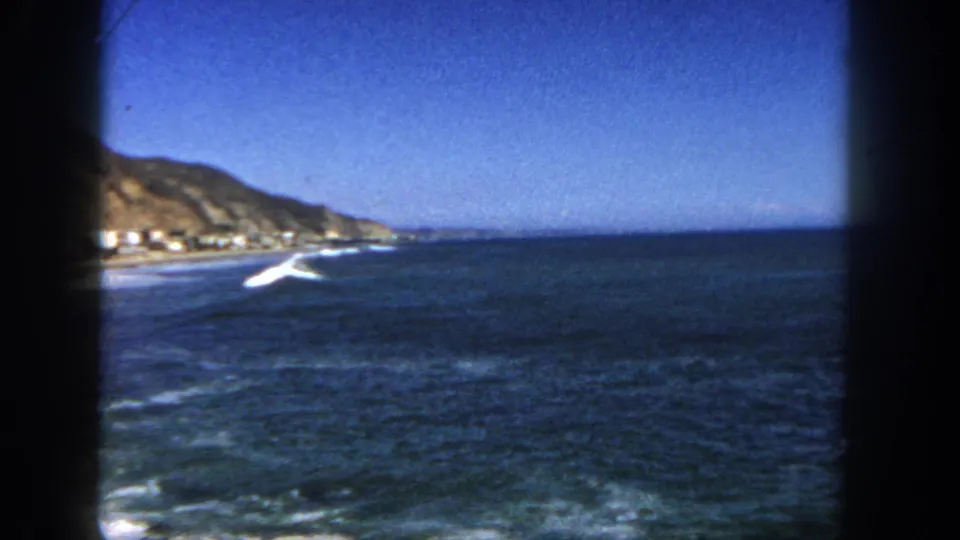 1962 San Diego coastal view showing open ocean, surf near shore, a distant cliff, blue sky, and visible 8mm frame edges.