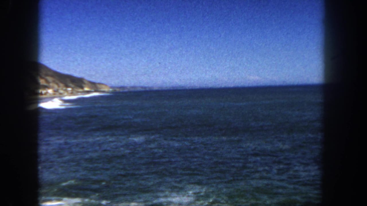 1962 San Diego coastal frame with a broad Pacific horizon, cliff edge at left, white surf, and saturated blue sky.