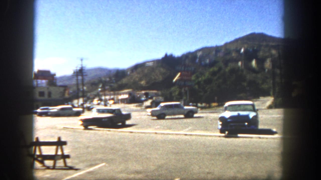 1962 San Diego street view with parked and moving cars, low buildings, utility poles, and hillside homes beyond the road.