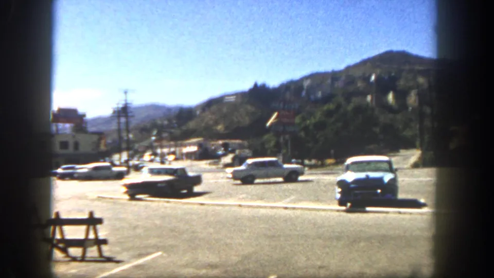 1962 San Diego street view with parked and moving cars, low buildings, utility poles, and hillside homes beyond the road.