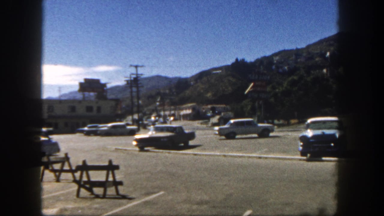 1962 San Diego roadside scene showing classic cars, utility poles, low buildings, hillside houses, and dark film-frame borders.