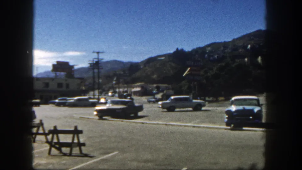 1962 San Diego 8mm frame with classic cars, roadside barricades, utility poles, and hillside homes behind a commercial street.