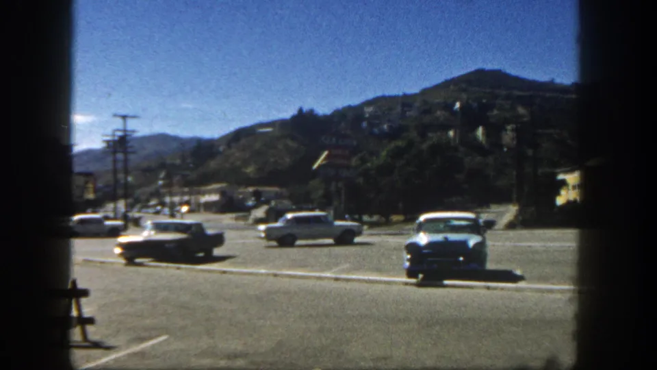 1962 San Diego street frame with a blue sedan, other classic cars, low roadside buildings, utility poles, and hillside homes.