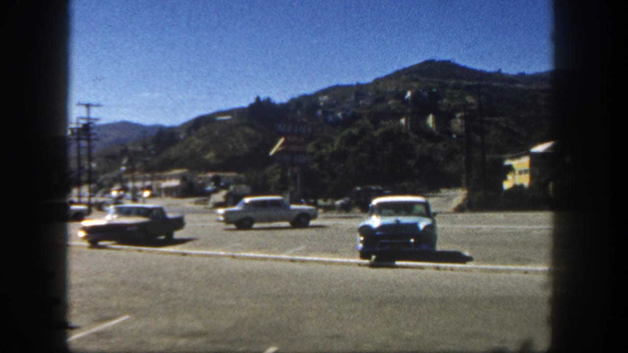 Classic cars in a sunny parking lot with a hillside behind them.
